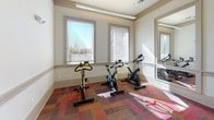 This secondary fitness center offers a dedicated space for cardiovascular workouts in a clean, well-maintained environment. The room features warm, neutral walls accented by classic crown molding and a chair rail, giving the space a polished, residential feel. Underfoot, a colorful patchwork carpet in rich tones of red, orange, and brown adds visual warmth while providing a comfortable exercise surface.

The fitness area is equipped with a variety of cardio machines, including treadmills, ellipticals, a recumbent bike with an interactive screen, and multiple upright spin bikes. Large windows fitted with horizontal blinds line one wall, allowing an abundance of natural light to fill the room while offering views of the surrounding outdoor area. A full-length mirror along one wall visually expands the space and aids users during workouts.

Two ceiling fans with pendant lighting help maintain air circulation and comfort throughout exercise sessions. A set of double panel doors provides access to an adjacent storage or utility area. The main entrance consists of double glass doors, framed with a posted notice board, ensuring easy access and clear communication for users. The overall ambiance is bright, functional, and inviting, making it a practical and comfortable fitness resource.