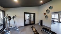 This single-floor fitness space is thoughtfully designed to deliver both functionality and modern aesthetic appeal. The workout room serves as the centerpiece of the space, characterized by crisp white walls that contrast sharply with a bold, dark ceiling — a striking design choice that lends the room a sleek, contemporary atmosphere. Recessed lighting is embedded throughout the dark ceiling, providing clean, even illumination across the entire exercise area. The flooring is finished in a smooth, neutral-toned material that complements the overall color palette and is well-suited to high-activity use. A set of black-framed French doors connects the fitness room to an adjoining area, allowing natural light to filter through while maintaining a visual separation of spaces. Exercise equipment, including a treadmill and a workout bench, is positioned throughout the room, making efficient use of the available floor space. On the wall, a set of geometric hexagonal shelving units adds a decorative yet practical touch, offering display space that enhances the room's modern character. A sleek, freestanding air purifier further speaks to the attention to comfort and wellness within the space. Overall, the design strikes a purposeful balance between a motivating workout environment and refined, contemporary style.