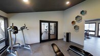 This single-floor fitness space is thoughtfully designed to deliver both functionality and modern aesthetic appeal. The workout room serves as the centerpiece of the space, characterized by crisp white walls that contrast sharply with a bold, dark ceiling — a striking design choice that lends the room a sleek, contemporary atmosphere. Recessed lighting is embedded throughout the dark ceiling, providing clean, even illumination across the entire exercise area. The flooring is finished in a smooth, neutral-toned material that complements the overall color palette and is well-suited to high-activity use. A set of black-framed French doors connects the fitness room to an adjoining area, allowing natural light to filter through while maintaining a visual separation of spaces. Exercise equipment, including a treadmill and a workout bench, is positioned throughout the room, making efficient use of the available floor space. On the wall, a set of geometric hexagonal shelving units adds a decorative yet practical touch, offering display space that enhances the room's modern character. A sleek, freestanding air purifier further speaks to the attention to comfort and wellness within the space. Overall, the design strikes a purposeful balance between a motivating workout environment and refined, contemporary style.