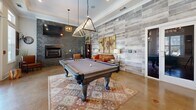 Located at 1709 Cason Ln in Murfreesboro, TN, this single-floor community lounge and billiards room offers a well-appointed gathering space with a thoughtfully designed interior. Polished concrete floors extend throughout, giving the space a sleek, modern foundation that complements the room's eclectic mix of rustic and contemporary design elements.

At the heart of the room sits a full-sized billiards table, illuminated by a striking geometric pendant light that serves as a focal point. One wall is clad in stacked stone, anchoring a built-in fireplace flanked by a wall-mounted television — creating a cozy seating area with a leather armchair and sofa. An adjacent accent wall features weathered wood-look tile, adding warmth and texture to the space.

Large double-pane windows dressed with white blinds flood the interior with natural light and offer views of the outdoor courtyard. A glass-paned double door provides direct access to the exterior. Crown molding and tall ceilings enhance the sense of openness throughout.

A dining nook near the windows features a round dark-wood table with industrial-style stools, ideal for casual seating. Decorative touches — including vintage shutters, potted greenery, and layered area rugs — complete the inviting, well-balanced atmosphere of this versatile community space.
