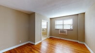 This single-floor space offers a practical and comfortable layout designed for everyday living. The main living areas are finished with rich, warm-toned hardwood flooring that adds character and continuity throughout the space. Neutral taupe-colored walls complement the light-toned textured ceilings, creating a cohesive and understated aesthetic.

The living area is well-lit by a large window fitted with horizontal blinds, allowing natural light to flow in while maintaining privacy. A wall-mounted air conditioning unit provides climate control directly within the room. An open doorway connects the living space to an adjacent area, enhancing the sense of flow and connectivity throughout the unit.

The kitchen is efficiently designed, featuring rich cherry wood cabinetry — both upper and lower — paired with granite countertops in warm yellow and grey tones. A stainless steel single-basin sink and a black electric range with an overhead exhaust hood round out the functional cooking space. Light-toned ceramic tile flooring adds a clean contrast to the kitchen's warm wood tones.

A separate tiled room — likely a dining area or secondary space — also features a window with blinds and a wall-mounted AC unit, and opens directly into the hardwood-floored living area. The front entry is defined by a clean white door, grounding the layout and offering a clear sense of arrival.
