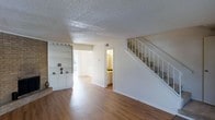 This two-story home offers a functional and straightforward layout designed for comfortable everyday living. The main floor features warm hardwood-style flooring that flows throughout the open living area, creating a cohesive and inviting atmosphere. A set of built-in shelves and cabinetry along one wall provides ample storage and display space, adding a practical yet classic architectural detail to the room. The living area opens toward a secondary room or dining space, bathed in natural light from an exterior window, enhancing the sense of openness. A glimpse into the adjacent kitchen reveals warm wood-toned cabinetry and a well-organized layout, making it a functional workspace. A staircase with white railings leads to the upper floor, where additional living or sleeping quarters are located. The textured ceiling adds a traditional finish to the interior throughout. Overall, the main floor presents a clean, neutral palette with white walls and trim that allows for versatile interior styling. The two-floor configuration provides a natural separation between the living spaces on the main level and the private areas above, making this home well-suited for a variety of living arrangements.