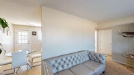 This single-floor residence offers a practical and well-organized layout across several distinct living spaces. The kitchen is equipped with dark cabinetry providing ample storage, granite-look countertops, white appliances including a refrigerator, electric range, microwave, and dishwasher, all set atop light tile flooring. The space opens toward a dining area, creating a connected and functional flow for daily living.

The living room features carpet flooring and is a generously sized area, accommodating a tufted leather sofa and glass-top coffee table. A mirrored display cabinet and indoor plants add character to the room, while natural light filters in through window blinds. A partial dividing wall separates the living area from the adjoining dining space.

The home includes multiple bedrooms, each finished with carpet flooring and textured ceilings. The primary bedroom features dual windows that allow for ample natural light and includes a closet with bifold doors. A secondary bedroom, suited for a younger occupant, also benefits from multiple windows and neutral wall tones. Throughout the home, textured ceilings and neutral color palettes contribute to a clean, understated aesthetic.