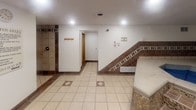 This single-floor spa and wellness facility offers a well-appointed retreat designed for relaxation and recreation. The layout encompasses several distinct functional areas that work together to create a comprehensive wellness experience.

At the heart of the facility is a generously sized hot tub, elevated on a multi-step stone and tile platform accented with decorative mosaic tile trim. Entry is made accessible via a metal safety handrail, and the surrounding floor features light-toned ceramic tile with diamond inlay accents throughout.

Adjacent to the hot tub area is a dedicated sauna room, enclosed with a glass-paneled door and fitted with natural wood benches and slatted flooring, offering an authentic dry-heat experience. Posted rules and signage reinforce safety and proper usage guidelines throughout the space.

A spacious transitional area connects the wet zones, featuring coat hooks, recessed lighting, and a clean tiled floor with consistent brown tile border accents along the base of the walls. This area also provides access to a separate lounge room, which includes a small café-style bistro table and chairs with striped cushions, a wall-mounted television, and a large interior window overlooking an adjacent room. The overall ambiance is functional and inviting, with recessed lighting providing consistent illumination throughout all areas of the facility.