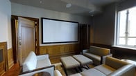 This elegant private home theater is designed to deliver a premium cinematic experience within a refined and comfortable setting. The room is anchored by a large, wall-mounted projection screen framed in sleek black, positioned above a wainscoted wall clad in warm, rich hardwood paneling that adds a classic and distinguished character to the space. The walls are finished in a muted gray tone that complements the dark wood accents and enhances the viewing environment by minimizing light reflection. Seating is generously appointed with plush, light-colored upholstered armchairs and cushioned ottomans arranged atop a patterned area rug, creating an inviting and relaxed atmosphere ideal for extended viewing sessions. The ceiling features recessed in-ceiling speakers seamlessly integrated into the design, ensuring high-quality audio without visual disruption. Subtle recessed lighting provides ambient illumination while maintaining the theater's immersive ambiance. Warm wood-trimmed doorways frame both sides of the room, adding architectural depth and continuity with the overall design aesthetic. The result is a thoughtfully designed space that balances functionality with luxury, offering both exceptional audiovisual performance and sophisticated interior style.