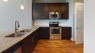 This single-floor space showcases a contemporary design with a cohesive blend of dark espresso cabinetry, granite countertops, and warm hardwood flooring throughout the main living areas. The kitchen serves as a central highlight, featuring flat-panel dark cabinetry extending to the ceiling, a classic white subway tile backsplash, stainless steel appliances including a range, built-in microwave, and a double-basin sink. A peninsula island with granite countertops provides additional workspace and casual seating, illuminated by stylish pendant lighting that adds warmth to the space.

The open layout connects the kitchen seamlessly to the living areas, creating an airy, functional flow. Bedroom spaces offer plush carpeting or hardwood flooring depending on the room, each equipped with ceiling fans and generously sized windows that allow natural light to fill the space. The neutral gray walls provide a versatile backdrop throughout.

The bathroom is finished with large-format tile flooring, full tile surround in the tub/shower combo, a dark vanity with granite countertop, and brushed nickel fixtures, maintaining the consistent modern aesthetic found throughout the unit.