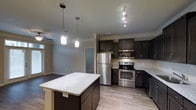 This single-floor residence showcases a thoughtfully designed open-concept layout that seamlessly connects the kitchen and living areas. The kitchen is a standout feature, anchored by a spacious central island topped with light granite countertops that provide ample prep and gathering space. Dark espresso cabinetry lines the walls, offering generous storage while creating a rich contrast against the light-toned tile backsplash and neutral walls. Stainless steel appliances — including a refrigerator, range, microwave, and dishwasher — complete the modern kitchen aesthetic. Track lighting above the cooking area and elegant pendant lights suspended over the island provide layered illumination throughout the space. Light hardwood-style flooring runs throughout, adding warmth and continuity as the space transitions from the kitchen into the adjacent living and dining areas. Crown molding details add a refined architectural touch to the ceilings. The open sightlines between rooms create an airy, connected feel, making the layout well-suited for both everyday living and entertaining. Overall, the space combines contemporary finishes with functional design for a clean, polished interior.