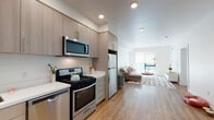 Located on 5th Street in Oakland's 94607 zip code, this single-floor unit showcases a thoughtfully designed, open-concept layout with a clean, contemporary aesthetic throughout. The entry leads directly into a galley-style kitchen equipped with stainless steel appliances, including a gas range, dishwasher, over-the-range microwave, and full-size refrigerator. Light wood-grain cabinetry lines the walls, complemented by white quartz-style countertops, providing ample storage and prep space. A striking red entry door adds a bold design accent to the otherwise neutral interior palette. Light hardwood flooring extends through the main living areas, connecting the kitchen seamlessly to the living room. The living area receives natural light through a large window and glass door that opens to an outdoor space, fitted with a roller shade for privacy. A dedicated home office features a horizontal clerestory window, offering a quiet and functional workspace. The bedroom includes carpeted flooring, a window with an exterior view, and sufficient room for comfortable furnishings. The bathroom is finished with stone-tile flooring, a wide vanity with undermount sink, and clean white walls. Recessed lighting is featured throughout, contributing to the bright and modern ambiance of the entire unit.