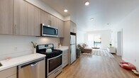 Located on 5th Street in Oakland's 94607 zip code, this single-floor unit showcases a thoughtfully designed, open-concept layout with a clean, contemporary aesthetic throughout. The entry leads directly into a galley-style kitchen equipped with stainless steel appliances, including a gas range, dishwasher, over-the-range microwave, and full-size refrigerator. Light wood-grain cabinetry lines the walls, complemented by white quartz-style countertops, providing ample storage and prep space. A striking red entry door adds a bold design accent to the otherwise neutral interior palette. Light hardwood flooring extends through the main living areas, connecting the kitchen seamlessly to the living room. The living area receives natural light through a large window and glass door that opens to an outdoor space, fitted with a roller shade for privacy. A dedicated home office features a horizontal clerestory window, offering a quiet and functional workspace. The bedroom includes carpeted flooring, a window with an exterior view, and sufficient room for comfortable furnishings. The bathroom is finished with stone-tile flooring, a wide vanity with undermount sink, and clean white walls. Recessed lighting is featured throughout, contributing to the bright and modern ambiance of the entire unit.