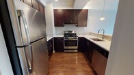 Located at 1031 John F. Kennedy Boulevard in Bayonne, New Jersey, this single-floor residence combines contemporary design with functional living. The open-concept layout connects the living area to the kitchen, creating a cohesive and welcoming environment. Rich hardwood flooring flows throughout the main living spaces, complementing the neutral-toned walls and recessed lighting overhead.

The kitchen is thoughtfully designed with dark espresso cabinetry, light quartz countertops, and a mosaic tile backsplash. Stainless steel appliances — including a gas range, refrigerator, and dishwasher — add a sleek, modern touch, while pendant lighting over the breakfast bar enhances both function and style.

The living area is bright and open, with large floor-to-ceiling windows that invite natural light and offer views of the surrounding neighborhood. A separate bedroom features plush carpeting and an expansive window with outdoor views, providing a comfortable and airy retreat.

The bathroom is clean and modern, featuring white subway tile surrounding a soaking tub and shower combination, a dark vanity with quartz countertop, a large mirror, and neutral large-format floor tiles. The overall design is cohesive, minimal, and move-in ready, making this a well-rounded residence in a convenient Bayonne location.