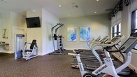 This fitness center offers a clean, well-organized space designed to support a variety of workout routines. The room features a durable carpet flooring suited for exercise, with recessed lighting throughout that ensures the space is consistently well-lit. A wall-mounted flat-screen television provides entertainment during workouts, adding to the overall comfort of the environment. The cardio section is equipped with spinning bikes and treadmills, catering to a range of aerobic exercise preferences. A multi-use strength training tower with weight stacks and an adjustable bench round out the strength training offerings. The neutral, light-toned walls are accented by two vibrant abstract art pieces in shades of blue and green, lending a fresh and energizing ambiance to the space. Ventilation is provided via ceiling vents, maintaining air circulation throughout workouts. The overall layout is thoughtfully arranged to maximize usability while maintaining an open, uncluttered feel. This fitness center provides a comprehensive and inviting environment suitable for both casual exercisers and those with more dedicated fitness routines.