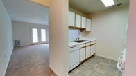 Located at 232 W. Gilbert St. in Hampton, Virginia, this single-story residence offers a straightforward and functional layout designed for comfortable everyday living. The kitchen is efficiently arranged with ample white cabinetry providing generous storage above and below the countertops. A double-basin sink sits beneath a clean white countertop, accompanied by a built-in dishwasher. The tile flooring in the kitchen is durable and easy to maintain, providing a practical contrast to the adjoining living area. A partial wall separates the kitchen from the rest of the space, creating a sense of definition while maintaining an open, connected feel. The living area flows naturally from the kitchen and features wall-to-wall carpeting that adds warmth underfoot. A set of French doors at the far end of the living space fills the room with natural light and offers an inviting connection to the outdoors. The neutral wall tones throughout the space create a clean, versatile backdrop that is easy to personalize. Overall, the residence presents a well-organized, single-level floor plan that balances practicality with a bright, airy atmosphere.