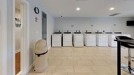 This single-story communal laundry facility is designed for practical, high-capacity use. The main laundry area is spacious and bright, finished with large-format light beige ceramic tile flooring and neutral gray walls that create a clean, functional atmosphere. Natural light filters in through windows fitted with white plantation shutters flanking a glass-paned entry door, which also provides views of the surrounding outdoor greenery.

The facility is equipped with multiple front-loading washing machines and top-loading dryers, all in white, arranged along the walls to maximize floor space and ease of circulation. A wall-mounted payment or card station is positioned near the entrance for convenient access, alongside instructional signage to assist users. A wall-mounted flat-screen television provides entertainment while waiting.

A secondary area connects through interior doors and features additional utility infrastructure, including an electrical panel, a wall clock, and a waste receptacle station with laundry supplies nearby. Security is supported by a ceiling-mounted camera. The space maintains a tidy, organized appearance with clear no-smoking signage and well-marked exits, ensuring a safe and user-friendly environment throughout.