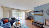 This single-floor residence offers a thoughtfully arranged layout that balances functionality and comfort throughout its living spaces. The open-concept living and dining area serves as the heart of the home, featuring warm hardwood-style flooring that extends throughout the main areas. The living room is bathed in natural light from large sliding glass doors fitted with vertical blinds, which provide access to an outdoor area framed by lush greenery. A bold blue accent wall defines the dining space, where a glass-top round table sits beneath decorative wall art, with a mirror that reflects the adjacent kitchen. The kitchen is equipped with warm oak cabinetry, dark granite-style countertops, a gas range, and a stainless steel sink, offering practical workspace for cooking. A front entry area provides a welcoming transition into the main living space. The primary bedroom features a striking terracotta-orange accent wall, carpet flooring, and a spacious closet with a full-length hanging rod. The second bedroom is finished in neutral tones with plush carpet and offers a calm, restful atmosphere. The bathroom includes white subway tile wainscoting, a vanity with wood cabinetry, and a tub-shower combination.