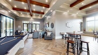 Located at 355 The Preserve Dr in Athens, Georgia, this single-floor clubhouse offers a thoughtfully designed communal space that blends comfort with modern style. The interior is anchored by striking herringbone-patterned wood-look tile flooring that flows seamlessly throughout, creating a cohesive and refined aesthetic. Exposed dark wood ceiling beams set within coffered and tray ceiling details add warmth and architectural character to the main lounge area, which is furnished with plush leather sofas and armchairs arranged around a wall-mounted flat-screen television. Large glass-paned French doors open to an outdoor pool area, flooding the space with natural light and providing easy indoor-outdoor connectivity. A dedicated game zone features a full-sized billiards table, while a casual dining and lounge area offers high-top tables and bar-height seating beneath pendant lighting, ideal for social gatherings. Oversized windows throughout the café-style nook frame views of the surrounding neighborhood and greenery. The neutral gray walls paired with curated décor, including decorative wall art, mirrors, and potted plants, contribute to an inviting and polished ambiance suited for both relaxation and recreation.