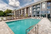 Located at 4781 SW 36 Rd in Gainesville, Florida, this outdoor amenity area showcases a resort-style pool deck designed for relaxation and recreation. The centerpiece is a generously sized, rectangular swimming pool with crystal-clear turquoise water, accessible via stainless steel entry rails and featuring shallow wading ledges along one side — ideal for lounging in the water. Surrounding the pool is a spacious stone-paved deck finished in a neutral travertine-style tile, providing ample room for residents to spread out and unwind. Dozens of adjustable chaise lounge chairs are arranged along the pool's perimeter, complemented by shade umbrellas in a signature light blue tone and occasional hanging egg chairs that add a relaxed, resort ambiance. Seating areas with outdoor dining furniture are also available for casual gatherings. The deck is enclosed by a black metal fence for safety, while lush mature trees beyond the perimeter provide a natural green backdrop. The multi-story residential building rises directly adjacent, with private balconies overlooking the pool area. Additional recreational features, including what appears to be a play area and hammock space, are visible nearby. The overall atmosphere is bright, open, and inviting, reflecting a modern community lifestyle under the sunny Florida sky.