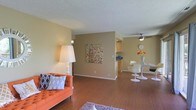 This single-floor space showcases an open-concept layout that seamlessly connects the living, dining, and kitchen areas into one cohesive environment. The living area is anchored by warm hardwood-style flooring that extends throughout, complemented by neutral beige walls that create a bright, welcoming atmosphere. A textured ceiling adds a classic architectural detail overhead.

The living space offers ample room for seating arrangements, as illustrated by the vibrant orange sofa that adds a pop of color against the understated backdrop. A tall floor lamp provides warm ambient lighting, enhancing the inviting feel of the room.

Toward the back, the dining area is defined by a sleek, modern round table surrounded by white tulip-style chairs, beneath a ceiling fan that aids in air circulation. A vase of fresh flowers on the table adds a lively touch.

The kitchen is partially visible beyond the central dividing wall, featuring cabinetry and countertops that maintain the space's clean, functional aesthetic. Overall, the open flow, consistent flooring, and neutral palette give this space a cohesive, comfortable, and well-organized character.