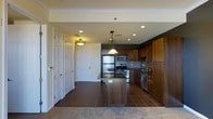This single-floor residence offers a well-designed open-concept layout that seamlessly connects the kitchen and living areas. The kitchen stands as the centerpiece of the space, showcasing warm medium-toned wood cabinetry that extends to the upper walls, complemented by a sleek stone-surface island that provides additional prep space and casual seating. A full suite of stainless steel appliances — including a refrigerator, range, microwave, and dishwasher — adds a modern, polished feel, while the tile backsplash introduces a subtle textural contrast. A decorative pendant light hangs above the island, adding an elegant focal point to the room. Dark ceramic floor tiles run throughout the kitchen and entry area, offering a rich, grounded aesthetic, while carpeting transitions into the adjacent living space for added comfort. Recessed lighting across the ceiling provides even, practical illumination throughout. The entryway is flanked by closet doors, offering generous storage right from the front door. Crown molding along the ceiling adds a refined architectural touch, elevating the overall finish of the space. The open layout and cohesive material palette create a functional yet inviting atmosphere throughout.