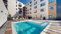 Located at 1060 Bolivar Road in Cleveland, Ohio, this generously sized outdoor amenity space is thoughtfully designed to serve as a vibrant social hub. The courtyard is framed by a modern multi-story building with a contemporary façade featuring large windows, wood-accented balconies, and a clean mix of white paneling and bold blue cladding that creates a striking architectural backdrop.

At the heart of the space is a sparkling outdoor swimming pool equipped with an accessibility lift, surrounded by a wood-deck pool surround and an expansive tiled deck lined with lounge chairs and Adirondack seating. A pergola structure at one end of the pool area provides a shaded retreat.

Adjacent to the pool, a large open-air patio features large-format tile pavers and is furnished with multiple dining table and chair sets, centered around a built-in stone grilling station — ideal for outdoor gatherings. String lights are strung overhead, creating an inviting atmosphere for evening use.

A vibrant botanical mural depicting maple leaves serves as an eye-catching artistic focal point alongside a section of lush artificial turf, perfect for lawn games such as cornhole. A dedicated lounge zone offers plush seating with colorful accent pillows, complemented by raised planters with greenery throughout. The entire space is enclosed by sleek black metal railing, contributing to its polished, modern aesthetic.