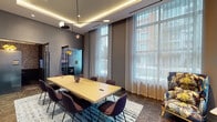 This thoughtfully designed single-floor workspace delivers a polished, contemporary environment suited for both collaborative and focused work. The centerpiece is a spacious conference room anchored by a large light-wood table surrounded by upholstered chairs in a rich plum hue. Floor-to-ceiling windows dressed with sheer white curtains flood the room with natural light and offer views of the surrounding urban streetscape. A sculptural pendant light fixture and a coved, LED-lit ceiling add architectural interest overhead, while dark stone-tile flooring and a patterned area rug ground the space with texture and warmth. A built-in credenza along one wall provides additional utility, accompanied by a wall-mounted display screen ideal for presentations. Complementing the main conference area is a vibrant tropical-print sofa, introducing an energetic design accent. Tucked away is an intimate private meeting pod, outfitted with a compact table, modern chairs, textured wallcovering, framed artwork, and a mounted flat-screen display — perfect for focused one-on-one sessions or video calls. A connecting corridor, lined with large-scale curated artwork and illuminated by recessed lighting, ties the spaces together with a gallery-like quality, reinforcing the space's commitment to a refined, professionally inspired atmosphere.