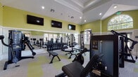 This fitness center is a comprehensively outfitted facility designed to support a full range of exercise routines. The space is divided into distinct zones, with cardio equipment — including treadmills and stationary bikes — positioned near large, multi-pane windows that flood the area with natural light and offer views of the surrounding outdoor environment. The bright, airy feel is enhanced by soaring ceilings with recessed lighting and a tray ceiling detail in the central area.

The strength training zone is well-appointed, featuring a full rack of dumbbells, adjustable workout benches, cable machines, and dedicated weight machines such as a leg extension unit. A floor-to-ceiling mirror runs along one wall, visually expanding the space and providing users with proper form feedback during workouts. Multiple wall-mounted flat-screen televisions are positioned throughout for entertainment.

The interior design is characterized by a two-tone wall treatment — a crisp white wainscoting base paired with a vibrant chartreuse-green upper wall — lending the space an energetic and modern aesthetic. A patterned carpet with green accent tiles completes the look. Glass-paneled doors connect to an adjacent common area, and a dedicated entry with posted fitness rules ensures an organized, community-oriented environment.