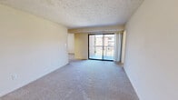Located at 7509 Mandan Rd, Unit 202 in Greenbelt, Maryland, this two-bedroom, one-bathroom residence offers a practical and comfortable single-floor layout. The spacious living room is a standout feature, finished with plush wall-to-wall carpeting and a textured ceiling. Large sliding glass doors open directly onto a private balcony, flooding the space with natural light and providing outdoor views of the surrounding neighborhood. An adjacent dining area connects seamlessly to a kitchen equipped with warm wood-toned cabinetry and dark countertops, visible through an open pass-through. The generously sized primary bedroom features a wide window with blinds for natural light and privacy, along with a closet for ample storage. The second bedroom is similarly appointed with carpeted flooring, a window overlooking the exterior, and its own closet. Both bedrooms are accessible via a connecting hallway that also leads to the full bathroom, which includes a tub/shower combo with clean white tile surround. Throughout the unit, neutral warm tones and light-colored walls create a bright, cohesive ambiance. Two hallways provide logical flow and additional storage access, rounding out this well-organized and functional home.
