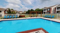This inviting outdoor amenity space centers around a generously sized rectangular swimming pool with vivid blue water and a classic brick coping border. The pool area is designed for both relaxation and recreation, offering ample open deck space finished in smooth concrete that surrounds the pool on multiple sides. Several blue lounge chairs are positioned along the perimeter, providing comfortable spots to unwind poolside.

A standout feature of the space is a natural wood pergola situated at the far end of the deck, adding a warm, architectural focal point and a shaded gathering area. Nearby, a cozy outdoor seating arrangement with red cushioned furniture offers an additional social space. The entire area is enclosed by a clean white aluminum fence, providing both safety and a defined boundary.

Beyond the fence, mature trees and well-maintained green lawns create a pleasant natural backdrop, while neighboring residential buildings are visible in the distance, lending a community-oriented atmosphere. The open-sky setting allows for abundant natural light throughout the day, enhancing the bright, welcoming character of the space. Overall, this pool area is thoughtfully designed to serve as a premier outdoor recreational and social amenity.