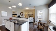 Located at 2000 Birchwood Circle in Bethlehem, Pennsylvania, this single-floor residence offers a thoughtfully designed open-concept living space that seamlessly connects the kitchen, dining, and living areas. The kitchen serves as a central hub, featuring dark granite countertops, white cabinetry, a stainless steel sink, a dishwasher, and a kitchen island with pendant lighting overhead — creating a clean, contemporary aesthetic. Rich hardwood-style flooring runs throughout, adding warmth and continuity to the space. The dining area comfortably accommodates a bistro-style table and chairs, positioned conveniently near a glass door that opens to a private balcony, allowing for natural light and outdoor views. The living area is open and inviting, providing ample room for comfortable seating arrangements. A decorative mirror and framed wall art contribute to the polished, cohesive interior design. A bedroom is visible through the open layout, offering a sense of the full living experience this residence provides. The neutral color palette of warm grays and whites throughout the space creates a bright, modern ambiance that feels both functional and welcoming, making it well-suited for everyday living.