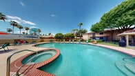 This single-story outdoor amenity space centers around a generously sized, freeform swimming pool with crystal-clear turquoise water, framed by a classic red brick coping that lends a warm, resort-inspired aesthetic. Adjoining the main pool is a circular spa, seamlessly integrated into the pool deck and finished with matching brick tile detailing. The expansive concrete sun deck surrounding the pool is outfitted with colorful lounge chairs and shaded seating areas featuring patio tables and large market umbrellas in vibrant red, blue, and green, ideal for relaxing under the open sky. The adjacent single-story clubhouse building, identified as unit 4401, showcases a distinctive Spanish-style architecture with terracotta clay tile roofing and smooth stucco exterior walls in a warm sand finish. Desert-friendly landscaping surrounds the structure, incorporating ornamental gravel beds, native rocks, and low-water plants such as fan palms and flowering shrubs. Mature palm trees and shade trees punctuate the grounds, providing natural canopy and a lush backdrop. A perimeter block wall with iron fencing ensures privacy while maintaining an open, airy feel. The entire space benefits from abundant natural sunlight and wide-open sky views, creating a bright and inviting outdoor retreat well-suited for leisure and community gatherings.