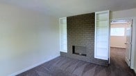 This single-story space centers around a commanding floor-to-ceiling brick fireplace that serves as the defining architectural feature of the living area. The fireplace is constructed of painted brick in a neutral gray-brown tone, extending the full height of the wall and creating a strong visual anchor for the room. Flanking the fireplace on both sides are symmetrical built-in shelving units finished in crisp white, providing ample display and storage opportunities while framing the hearth in a balanced, classic arrangement. The raised brick hearth extends outward at the base, adding depth and dimension to the feature wall. The flooring throughout is wall-to-wall carpet in a medium gray-brown tone, contributing to a warm and comfortable underfoot feel. The walls are painted in a clean, light neutral that reflects available light and lends an open, airy quality to the space. Smooth, white baseboards run along the perimeter, complementing the built-in shelving and providing a cohesive finish. The overall ambiance is cozy yet functional, with the fireplace and built-ins offering both character and practicality suited to a comfortable living environment.