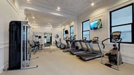 This fitness facility masterfully merges historic architectural elegance with contemporary workout amenities across a single, generously proportioned floor. The main gym area is defined by its striking ornate plaster ceiling with intricate molding and medallion details, painted crisp white and fitted with recessed lighting that illuminates the space evenly. Multiple black-framed windows line the walls, drawing in natural light and providing views of the surrounding urban streetscape. The cardio zone is well-equipped with a row of treadmills and elliptical machines, each positioned for optimal spacing, complemented by wall-mounted flat-screen televisions. A dedicated strength-training area features a multi-functional cable machine, free weights, and a bench. Full-length mirrors expand the sense of space throughout. Classical wainscoting and paneled wall detailing are consistent throughout all areas. A separate room with rich dark-wood millwork, gray carpeted flooring, and a large mirror serves as an additional training space. Adjoining the main gym is a serene yoga and stretching room featuring herringbone hardwood floors, sheer-draped windows on multiple walls, and a handsome dark marble fireplace with a mirrored surround, creating a calm and refined atmosphere for mindful movement.