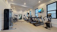 This fitness facility masterfully merges historic architectural elegance with contemporary workout amenities across a single, generously proportioned floor. The main gym area is defined by its striking ornate plaster ceiling with intricate molding and medallion details, painted crisp white and fitted with recessed lighting that illuminates the space evenly. Multiple black-framed windows line the walls, drawing in natural light and providing views of the surrounding urban streetscape. The cardio zone is well-equipped with a row of treadmills and elliptical machines, each positioned for optimal spacing, complemented by wall-mounted flat-screen televisions. A dedicated strength-training area features a multi-functional cable machine, free weights, and a bench. Full-length mirrors expand the sense of space throughout. Classical wainscoting and paneled wall detailing are consistent throughout all areas. A separate room with rich dark-wood millwork, gray carpeted flooring, and a large mirror serves as an additional training space. Adjoining the main gym is a serene yoga and stretching room featuring herringbone hardwood floors, sheer-draped windows on multiple walls, and a handsome dark marble fireplace with a mirrored surround, creating a calm and refined atmosphere for mindful movement.