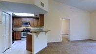 This single-floor residence showcases an open and airy layout designed for comfortable everyday living. The kitchen is thoughtfully appointed with warm wood cabinetry, granite countertops, and stainless steel appliances, including a range and built-in microwave. A skylight overhead floods the kitchen with abundant natural light, highlighting the clean tile flooring underfoot. A peninsula breakfast bar with corbel accents extends into the living space, offering a casual dining or gathering area that effortlessly connects the kitchen to the rest of the home. The vaulted ceilings rise dramatically, amplifying the sense of openness and volume throughout the main living area. Neutral-toned carpeting in the living and adjoining rooms creates a warm, cohesive feel as the space flows naturally from one area to the next. A wide doorway leads to an additional room, suggesting flexible use as a bedroom, office, or den. The overall design balances functionality with an inviting ambiance, making it a well-suited space for both daily routines and casual entertaining. Light, neutral wall tones throughout contribute to the home's bright and welcoming atmosphere.
