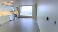 This modern high-rise unit offers a clean, open-concept layout that seamlessly blends the living and kitchen areas into a single, airy space. The standout feature is a large floor-to-ceiling window that floods the interior with abundant natural light while framing expansive panoramic city views, giving the space an elevated, urban feel.

The kitchen is positioned along one wall, featuring white cabinetry with a sleek, minimalist design, stainless steel appliances including a range and over-the-range microwave, and a light-toned countertop that complements the overall neutral color palette. The open living area benefits directly from the natural light pouring in through the oversized window, creating a bright and welcoming atmosphere.

Light-toned hardwood-style flooring runs throughout the space, contributing to the clean and contemporary aesthetic. The white walls and ceilings enhance the sense of openness and allow the cityscape backdrop to take center stage. The unit's high-floor positioning is evident from the bird's-eye view of the surrounding urban landscape visible through the window, adding a dynamic and distinctive character to everyday living. The overall ambiance is modern, minimal, and light-filled.