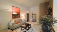 This single-floor space showcases a thoughtfully designed living area that blends modern aesthetics with a warm, inviting ambiance. The living room features neutral walls accented by a striking large-scale orange and brown artwork, which serves as a bold focal point. Elevated ceilings, enhanced by a ceiling fan, contribute to an open and airy atmosphere throughout the space.

The living area is furnished with a comfortable neutral-toned sofa accented with orange throw pillows, complementing the artwork and creating a cohesive color palette. A dark wood coffee table set rests atop a patterned area rug with circular designs, adding visual interest to the floor. A freestanding ladder-style bookshelf provides both storage and display space along one wall, while a lush indoor plant introduces a natural, organic element to the room.

A warm-toned accent wall frames a key architectural transition, adding depth and dimension to the interior. An open passage leads to an adjacent area, suggesting a well-connected floor plan. The overall design reflects a balance of contemporary style and functional living, with carefully curated décor that enhances the space's character.