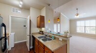 Located at 6805 Lebanon Rd in Frisco, TX, this single-floor residence offers a well-designed layout with a warm, neutral color palette throughout. The kitchen is a functional highlight, featuring rich wood cabinetry, granite-style countertops, a double stainless steel sink, and a breakfast bar peninsula ideal for casual dining. Stainless steel appliances—including a refrigerator, microwave, and range—are complemented by track lighting and distinctive amber pendant lights that add a touch of character. Dark hardwood-style flooring in the kitchen transitions to plush carpet in the living and bedroom areas. The living room impresses with soaring vaulted ceilings, crown molding, and a ceiling fan, creating an airy and open atmosphere. Multiple windows fitted with horizontal blinds allow ample natural light to fill each room. The bedrooms are generously sized with vaulted or elevated ceilings, ceiling fans, and multiple windows offering outdoor views. The bathroom includes a full tub-shower combination with large-format tile surround. Overall, the space delivers a comfortable and practical living environment with thoughtful design details throughout.