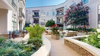 Located at 1180 Canton St in Roswell, Georgia, this stunning outdoor courtyard serves as the centerpiece of a well-appointed multi-story residential community. The space is thoughtfully designed to create a welcoming, resort-like atmosphere that encourages relaxation and social interaction.

At the heart of the courtyard stands a decorative circular fountain, surrounded by multiple seating arrangements featuring teak-framed furniture with plush white cushions. The seating areas are generously distributed throughout the space, offering both intimate and communal gathering spots ideal for residents and guests.

Curving brick-bordered planters wind through the courtyard, filled with a rich variety of lush shrubs, ornamental grasses, flowering hydrangeas, vibrant crepe myrtles, and mature shade trees. The layered landscaping adds depth and color throughout the seasons, enhancing the overall ambiance with natural beauty.

Wide, smooth concrete pathways meander through the greenery, providing easy access to all areas of the courtyard. Ceiling fans mounted on covered overhangs along the building's facade provide comfort during warm Georgia summers, while private balconies on the upper floors overlook the serene courtyard below.

The surrounding building features a classic yet contemporary exterior with gray horizontal siding, large multi-pane windows, and brick accents. The overall environment is tranquil, polished, and meticulously maintained, making this courtyard a standout amenity of the community.