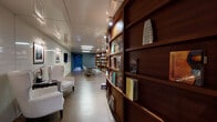 Located at Oasis Grand Boulevard in Fort Myers, Florida, this single-floor amenity space serves as a refined library and lounge area. The layout flows seamlessly from an intimate reading corridor into a broader open gathering area, creating a well-organized and functional environment.

A defining architectural feature is the curved, floor-to-ceiling built-in bookcase crafted from rich, warm-toned wood. Filled with an extensive and varied collection of books, this sweeping shelving unit anchors the space and lends it a scholarly yet welcoming character. Recessed spotlights integrated into the bookcase provide focused illumination, highlighting the collection and adding warmth to the interior.

On the opposing wall, high-gloss white paneling provides a clean, modern contrast. Gallery-style framed artwork — primarily nature and landscape photography — is displayed along this wall with individual picture lights, giving the space a polished, curated aesthetic. A flat-screen television is also mounted along this corridor, adding a media component to the lounge experience.

White wingback chairs and small side tables furnish the reading area, offering comfortable spots for quiet browsing. Toward the back, a sleek dark conference table surrounded by modern mesh chairs provides a dedicated workspace or meeting area. The flooring throughout is a neutral, matte finish that complements the overall contemporary design. Recessed linear ceiling lighting ensures the entire space is evenly and efficiently illuminated.