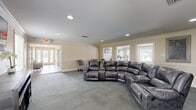 Located at 4950 US-90 W in Mobile, Alabama, this single-floor amenity space offers a thoughtfully designed environment that balances comfort and functionality. The interior is finished with warm, neutral tones, crown molding, chair-rail wainscoting, and plush carpet tiles throughout, lending a polished and welcoming atmosphere.

The living room serves as a social hub, featuring a large sectional recliner sofa and colorful accent chairs that invite relaxation and conversation. Recessed lighting and a window providing natural light enhance the open, airy feel of the room. A workstation area with dual computer monitors and white ergonomic chairs is conveniently positioned near a window, offering a practical space for residents to handle business or personal tasks.

The adjacent exercise room is well-equipped for fitness enthusiasts, housing a treadmill, elliptical machine, rowing machine, and a weight bench. Wall-mounted flat-screen TVs add entertainment options during workouts. Large French-style windows connect the exercise room visually to the living area, maintaining a sense of openness between the two spaces. Together, these rooms create a versatile, community-oriented facility designed to support both active and leisurely lifestyles.