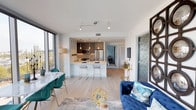 This contemporary single-floor residence is defined by its expansive floor-to-ceiling windows, which frame striking urban skyline and waterfront views while flooding the open-concept living spaces with natural light. The living and dining area flows seamlessly into a well-appointed kitchen featuring a large quartz waterfall island with bar seating, sleek wood-grain cabinetry, stainless steel appliances including a gas range, and elegant pendant lighting. Light hardwood flooring and clean white walls run throughout, creating a bright, cohesive aesthetic.

The two bedrooms each showcase bold, distinct design sensibilities. One features richly textured silver wallcovering, globe-style brass lighting fixtures, and a floor-to-ceiling window with city views. The second bedroom presents a serene atmosphere with a marble-effect accent wall, canopy-style drapery, and direct access to an outdoor balcony through floor-to-ceiling glass.

Both bathrooms are thoughtfully finished with marble-style tile flooring, dark cabinetry with illuminated mirrors, and walk-in closets. One bathroom includes a glass-enclosed shower, while the other offers a soaking tub with dark tile surround. The overall ambiance is polished and contemporary throughout.