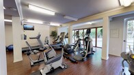 This fitness center is a thoughtfully designed, single-floor facility offering a full suite of exercise equipment to support a well-rounded workout routine. The space is organized into distinct zones, seamlessly connected by rich hardwood flooring that runs throughout. The cardio area is positioned along a wall of large floor-to-ceiling windows and glass doors, allowing abundant natural light to fill the room and providing open views of lush outdoor greenery. This section is equipped with multiple treadmills, elliptical trainers, and stationary recumbent bikes, each placed on protective rubber mats. A wall-mounted flat-screen television adds convenience for users during cardio sessions. The strength training area occupies a separate yet connected section of the facility, housing a multi-station cable machine, a functional trainer, and a cable row machine. Stability balls, medicine balls in varying weights, and additional accessories are neatly arranged along the walls, catering to flexibility and functional fitness needs. A large mirrored wall in the strength zone enhances the sense of space and allows users to monitor their form. An adjacent glass-enclosed room, visible through sleek black-framed glass partitions, appears to serve as a dedicated studio space. Overhead fluorescent lighting ensures the entire facility is well-lit, and crisp white walls with crown molding give the space a clean, polished appearance throughout.