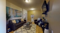 This single-floor unit offers a thoughtfully arranged layout designed for comfortable shared living. The welcoming living room sets a warm, inviting tone with rich hardwood-style flooring, bold gold-yellow accent walls, and crisp white ceilings. A dark leather sofa anchors the space alongside a cushioned armchair, coffee table, and a patterned area rug, all complemented by vibrant abstract artwork that adds a pop of color. The kitchen features rich wood cabinetry, granite-style countertops, a full suite of black appliances including a range, dishwasher, and microwave, and ample upper storage — offering everything needed for everyday meal preparation. The bedrooms are carpeted and designed for individual comfort, each accommodating a bed and dedicated desk workspace, with windows that invite natural light. The bathrooms are finished with light tile flooring, dark wood vanities with generous counter space, and tub-shower combinations, providing practical and clean shared facilities. The overall ambiance throughout the unit is bright and cheerful, with a cohesive color palette of teal, gold, and neutral tones that creates a lively yet functional environment well-suited for student or shared occupancy living.