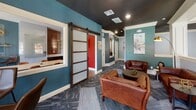 This thoughtfully designed single-floor communal space blends contemporary style with classic architectural details to create a welcoming and functional environment. The lounge area anchors the space with rich brown leather seating arranged around a low coffee table, set beneath a dramatic dark ceiling that contrasts elegantly with crisp white crown molding and wainscoting. Coffered ceiling details in the seating nook add a refined, traditional touch, while recessed lighting provides a warm ambient glow throughout.

Bold teal-painted walls flow through the space, creating a cohesive yet vibrant atmosphere. A rustic barn-style sliding door with frosted panel inserts serves as a standout architectural feature, dividing areas while adding an eclectic, industrial charm. Multiple windows throughout allow generous natural light and offer views of the surrounding neighborhood.

A dedicated meeting or conference area features a large rectangular table with leather chairs, a glass whiteboard wall, and a copper-toned brick tile accent wall with open glass shelving and dark cabinetry — ideal for collaborative work or presentations. A bar-height bistro table tucked near a corner window provides an additional casual seating option. A business center nook with a multifunction printer rounds out the practical amenities, making this space as functional as it is visually striking.