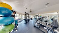 This fitness center offers a comprehensive workout facility thoughtfully organized across a single floor. The space is divided into two functional zones, seamlessly connected to provide an efficient and comfortable exercise environment.

The main workout area is bathed in natural light, courtesy of large multi-pane windows that line the walls and offer pleasant views of the surrounding landscaped grounds. The vaulted ceiling enhances the sense of openness, while a ceiling fan ensures comfortable airflow throughout. Recessed lighting supplements the abundant daylight for consistent illumination at all hours.

The cardio zone is well-appointed with multiple treadmills, elliptical trainers, a stationary bike, and a rowing machine, all arranged to maximize space and usability. A large floor-to-ceiling mirror along one wall visually expands the room and allows users to monitor their form. Wall-mounted televisions add entertainment options during workouts.

The strength training area features a multi-station cable machine, a full dumbbell rack with adjustable benches, medicine balls, stability balls, and various free weight accessories. Durable rubber flooring covers the entire space, providing safety and sound absorption. French doors offer convenient access to the exterior, rounding out a well-designed, fully functional fitness facility.