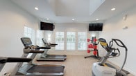 This single-story community facility is thoughtfully designed to offer residents both comfort and functionality across its distinct spaces. The main lounge area serves as the heart of the facility, anchored by a striking floor-to-ceiling stone fireplace with a rustic wood mantel and framed landscape artwork above. Plush seating, including cream sofas and wingback chairs in soft gray tones, is arranged to encourage conversation and relaxation. Tall ceilings and large French doors with gridded glass panes flood the space with natural light while providing direct access to an outdoor area. A multi-arm chandelier adds a refined touch overhead, complementing the neutral, sophisticated color palette throughout.

An adjacent seating area extends the lounge with additional sofa and dining arrangements beneath recessed lighting and another elegant chandelier, creating a versatile space for casual gatherings or individual use. The tile-accented entryway leads seamlessly into the carpeted common areas.

The dedicated fitness center is well-equipped with multiple Matrix treadmills, a Lateral X elliptical trainer, and a rack of weighted medicine balls. Wall-mounted flat-screen televisions and large windows with plantation shutters offering outdoor views keep the environment bright and engaging during workouts.