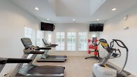 This single-story community facility is thoughtfully designed to offer residents both comfort and functionality across its distinct spaces. The main lounge area serves as the heart of the facility, anchored by a striking floor-to-ceiling stone fireplace with a rustic wood mantel and framed landscape artwork above. Plush seating, including cream sofas and wingback chairs in soft gray tones, is arranged to encourage conversation and relaxation. Tall ceilings and large French doors with gridded glass panes flood the space with natural light while providing direct access to an outdoor area. A multi-arm chandelier adds a refined touch overhead, complementing the neutral, sophisticated color palette throughout.

An adjacent seating area extends the lounge with additional sofa and dining arrangements beneath recessed lighting and another elegant chandelier, creating a versatile space for casual gatherings or individual use. The tile-accented entryway leads seamlessly into the carpeted common areas.

The dedicated fitness center is well-equipped with multiple Matrix treadmills, a Lateral X elliptical trainer, and a rack of weighted medicine balls. Wall-mounted flat-screen televisions and large windows with plantation shutters offering outdoor views keep the environment bright and engaging during workouts.
