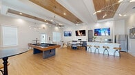 Located at 2401 Stanford Rd in Panama City, FL, this well-appointed single-story amenity space offers a thoughtfully designed interior that balances recreation, relaxation, and functionality. The open main area is anchored by light hardwood flooring and soaring ceilings adorned with warm wood beam accents, recessed lighting, and contemporary multi-arm pendant fixtures that lend a modern yet inviting ambiance. Large windows fitted with white horizontal blinds flood the space with natural light, enhancing the clean, bright aesthetic throughout.

The centerpiece of the main gathering area is a full-size pool table, complemented by a comfortable lounge seating arrangement with neutral-toned armchairs and a coffee table. A dedicated dining area with a table and chairs sits nearby, while multiple wall-mounted flat-screen TVs provide entertainment throughout. The kitchenette is well-equipped with dark wood cabinetry, white countertops, a stainless steel refrigerator, microwave, and a bar-height island with rustic wooden stools — ideal for social gatherings. The space also includes a private office, a closet, and two full bathrooms, making it a fully functional and versatile facility for its users.
