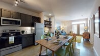 Located at 6710 Wahoo Drive in Myrtle Beach, SC, this single-story unit offers a thoughtfully designed open-concept layout that seamlessly connects the kitchen, dining, and living areas. The kitchen is well-equipped with dark shaker-style cabinetry, granite countertops, stainless steel appliances including a refrigerator, electric range, built-in microwave, and dishwasher. A built-in wine rack above the upper cabinets adds a distinctive touch, while polished concrete flooring lends an industrial-modern character throughout the main living areas. The dining space centers around a rustic wood farmhouse table with seating for six, positioned between the kitchen and the living room for easy flow. The living room features natural light from a set of large windows and a glass door, complemented by open wood shelving for display and storage. The bedroom offers a ceiling fan, multiple windows with blinds and curtains, and a warm, coastal-inspired aesthetic. The bathroom includes a shower-tub combo with overhead cabinet storage and polished concrete flooring. Completing the unit, a dedicated laundry room houses a full-size washer and dryer alongside a water heater. The overall ambiance blends coastal charm with modern finishes throughout.
