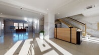 This refined building lobby makes a strong first impression through a combination of classic materials and contemporary design. Polished terrazzo floors extend throughout the space, reflecting abundant natural light that pours in through large, floor-to-ceiling glass entry doors framed in sleek black metal. Structural columns clad in richly veined marble anchor the open layout, complemented by marble wall paneling that lends a timeless elegance to the interior.

At the heart of the lobby, a warm natural wood reception desk with black metal accents serves as a welcoming focal point. A grand staircase with gleaming gold-toned railings rises prominently from the main floor, adding a touch of luxury and architectural drama. Recessed ceiling lighting, crown molding details, and crisp white walls enhance the sense of brightness and refinement throughout.

Adjacent to the reception area, a well-appointed lounge invites visitors to settle in, furnished with contemporary seating including a navy sofa, sculptural accent chairs, and a striped amber area rug. An arched floor lamp and potted greenery add warmth and life to the arrangement. A dedicated elevator bank, finished with stainless steel doors and warm mosaic tile surrounds, provides seamless vertical access. Together, these elements create a polished, professional, and welcoming environment.
