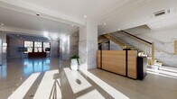 This refined building lobby makes a strong first impression through a combination of classic materials and contemporary design. Polished terrazzo floors extend throughout the space, reflecting abundant natural light that pours in through large, floor-to-ceiling glass entry doors framed in sleek black metal. Structural columns clad in richly veined marble anchor the open layout, complemented by marble wall paneling that lends a timeless elegance to the interior.

At the heart of the lobby, a warm natural wood reception desk with black metal accents serves as a welcoming focal point. A grand staircase with gleaming gold-toned railings rises prominently from the main floor, adding a touch of luxury and architectural drama. Recessed ceiling lighting, crown molding details, and crisp white walls enhance the sense of brightness and refinement throughout.

Adjacent to the reception area, a well-appointed lounge invites visitors to settle in, furnished with contemporary seating including a navy sofa, sculptural accent chairs, and a striped amber area rug. An arched floor lamp and potted greenery add warmth and life to the arrangement. A dedicated elevator bank, finished with stainless steel doors and warm mosaic tile surrounds, provides seamless vertical access. Together, these elements create a polished, professional, and welcoming environment.